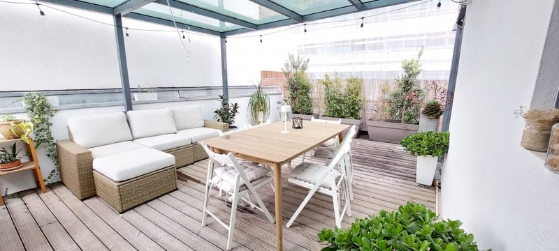 Covered terrace with a glass roof, a white outdoor sofa, a wooden dining table and planters