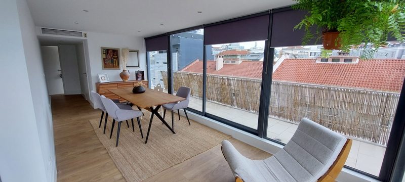 Lounge with a wooden dining table, a rocking chair and floor-to-ceiling windows over red-tiled rooftops